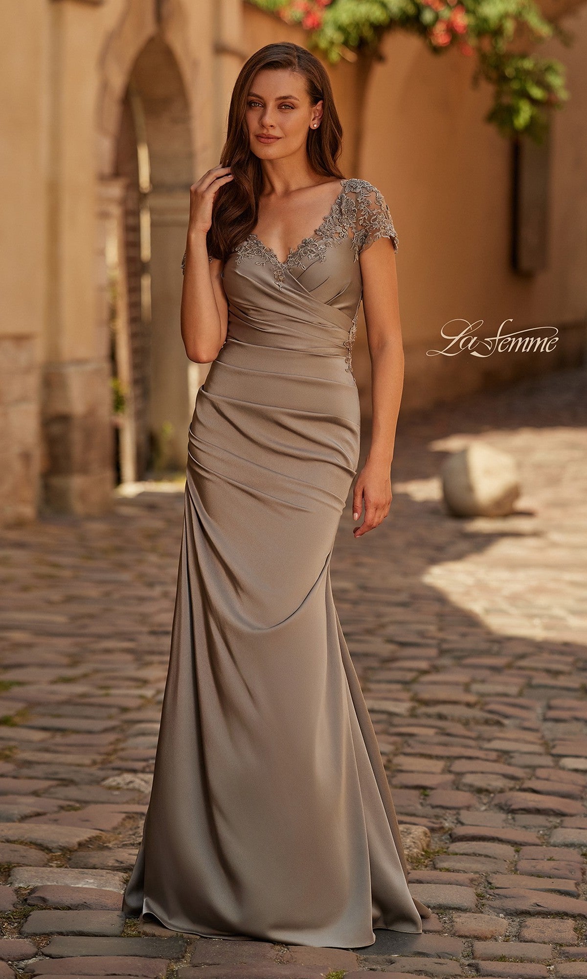 A woman stands on a cobblestone street wearing the La Femme 32431, a pewter long formal dress with a floor-length V-neck and beaded shoulder embellishments.